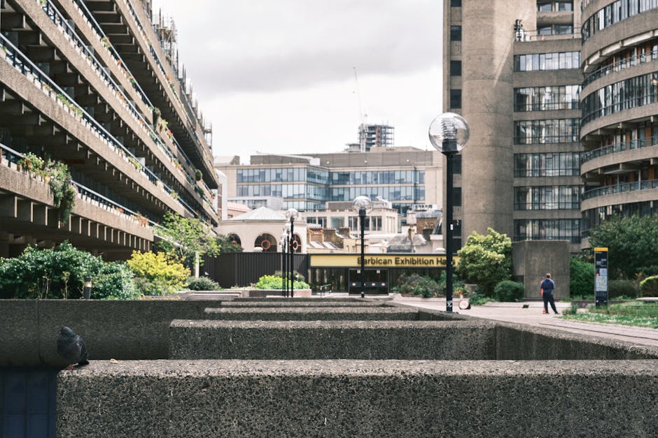 A wide shot of the exterior area of the Barbican Centre, showing a paved courtyard with series of rectangular concrete planters and steps in the foreground. Five black street lamps with spherical glas