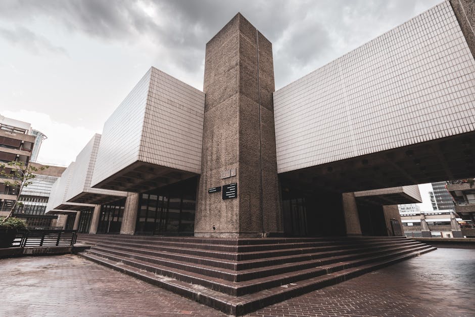 A wide shot of the exterior area of the Barbican Centre, showing a paved courtyard with series of rectangular concrete planters and steps in the foreground. Five black street lamps with spherical glas