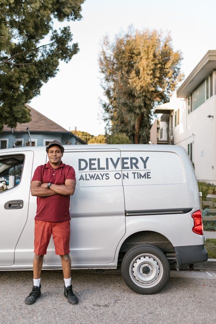 Two men are engaged in a home relocation process, loading a large cardboard box into the rear of a white delivery van parked on a brick driveway. The man on the left is wearing a black padded vest ove