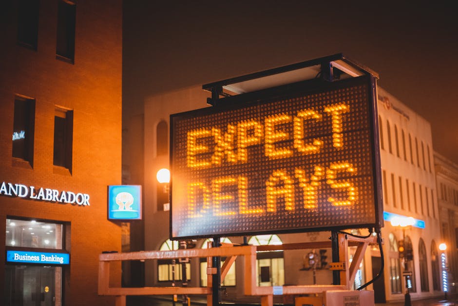 A large illuminated electronic road sign displaying the message 'EXPECT DELAYS' in bright yellow LED lights is positioned on a scaffolding structure along a city street at night. The background features a building with multiple windows, some of which are lit, and a sign for 'Land Labrador' on the brick facade. To the left of the electronic sign, there is a smaller blue-lit sign indicating 'Business Banking.' The street is illuminated by round street lamps, casting a warm glow over the scene. The scene suggests an urban environment experiencing traffic disruptions, which could impact home relocation or furniture transport services. The sign's message about delays is relevant in the context of city road access problems that may affect moving logistics, aligning with the page title 'City Road Removals Access Problems and Delays' on manandvanfinsbury.co.uk. This setting emphasizes the importance of planning for potential delays during house removals or moving operations in busy city areas.