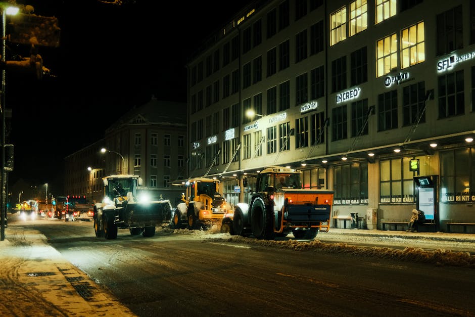 Nighttime view of a city street with three large yellow and black snowplough vehicles actively engaged in clearing snow from the road. The snow is being pushed aside onto the pavement along the curb. The street is illuminated by streetlights and the glow from nearby building windows, some of which display illuminated signage. The buildings lining the street are multi-story with large windows and modern façades. One building features signage for various companies, and a bench with a seated individual is visible outside. The snowploughs are positioned side by side, working to manage winter weather conditions, representing a typical urban scene during snow removal operations. This scene is relevant to house removals and moving logistics when considering winter weather challenges and the importance of clearing pathways for safe access, which is a service sometimes handled by companies like Man and Van Finsbury.