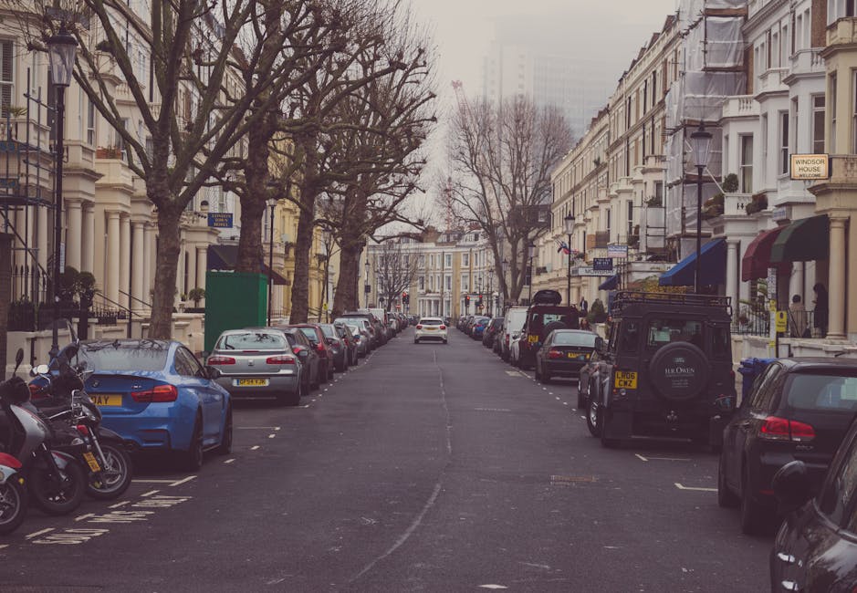 A straight residential street with parked cars lining both sides on a cloudy day. The vehicles include a blue sports car, several sedans, and an off-road style vehicle near the curb, with motorcycles parked in designated areas. Tall, ornate Victorian-style terraced houses with bay windows and decorative facades are visible on either side, some with awnings and potted plants on the balconies. Leafless trees cast shadows over the pavement, and street signs indicating parking restrictions and local notices are attached to poles. A small green utility box is situated on the sidewalk next to one of the trees, and the scene suggests an urban area suitable for home relocation or furniture transport. The overall environment depicts a typical London residential street, with a focus on parking arrangements and the urban setting associated with house removals.