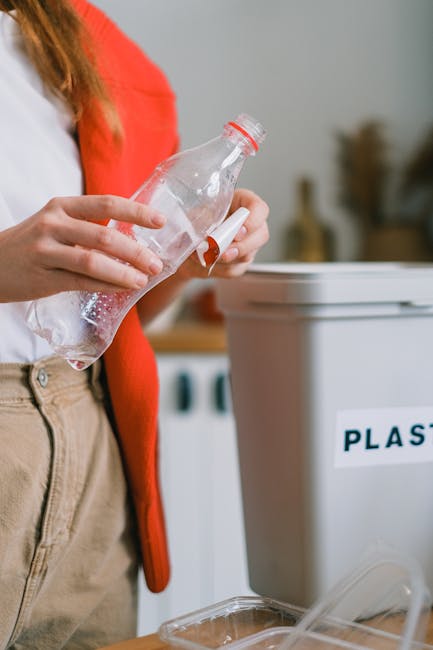 A person wearing a white shirt and beige trousers, with a red cardigan draped over their shoulder, is holding an empty plastic water bottle above a transparent plastic container. In the background, there is a white recycling bin with a label reading 'PLASTIC' on it, positioned near a kitchen or utility area. The scene suggests preparation or sorting of recycling materials, which may be part of the packing and organisation tasks involved during home relocation or furniture transport. The environment is well-lit with natural or ambient lighting, and the focus is on the hands and the plastic bottle, with background objects slightly blurred to emphasise sorting activity. This image relates to the logistics of moving and the importance of proper packing and disposal during house removals, as handled by services like Man and Van Finsbury.