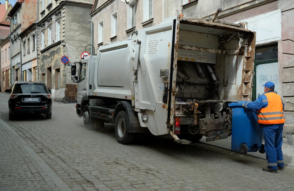 A municipal waste collection truck parked on a cobblestone residential street during daytime, with a sanitation worker dressed in a blue uniform and high-visibility orange vest standing beside the truck, holding the lid of a blue wheeled bin. The truck's rear hatch is open, revealing the empty mechanical compactor area used for collecting household rubbish, with some rust and debris visible inside. A black car is parked further along the narrow street, lined with old brick and plaster buildings with multiple windows. The scene depicts the loading process typical of local waste disposal services, with the truck positioned close to a doorway for efficient collection. The environment is well-lit with natural daylight, and the street has no other visible activity or pedestrians, emphasizing the routine operation of household waste collection and compliance with local disposal rules, relevant to house removals and clearance processes by [COMPANY_NAME].