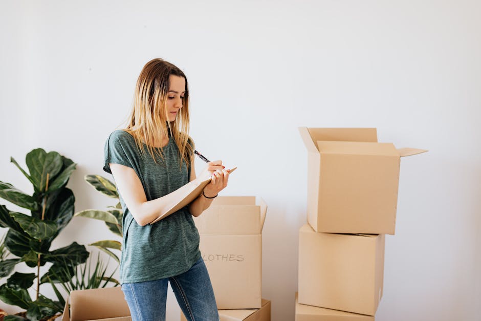A young woman with long blonde hair, dressed in a casual grey T-shirt and jeans, is standing indoors next to several packed cardboard boxes, some sealed and others open with flaps raised. She is writing on a notepad with a pen, likely making notes related to her home relocation or packing process. Behind her, a large green houseplant adds a natural element to the neutral white-wall setting. In the background, there are additional boxes stacked on the floor, with one partially visible labeled 'CLOTHES.' The scene captures a moment of organisation during packing or a residential move, aligning with the services offered by Man and Van Finsbury for small flat removals and furniture transport as part of comprehensive house removals and relocation logistics.