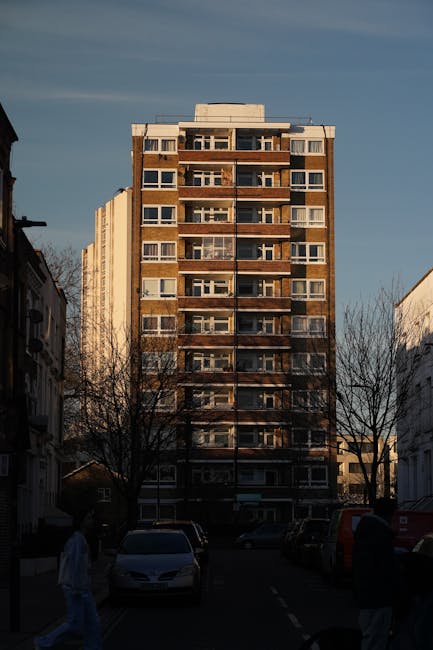 An exterior view of a multi-storey residential building on Golden Lane Estate, FINSBURY, shown during daytime with clear blue skies. The building features a brick façade with multiple balconies, large windows, and a flat roof. In the foreground, the street is visible with several parked cars along the pavement and two pedestrians walking past. Bare trees line the street, indicating a possible late autumn or winter season. The scene represents an urban residential area, relevant to home relocations and furniture transport, as part of the packing and moving process conducted by companies like Man and Van Finsbury, specialising in removals services within the area.