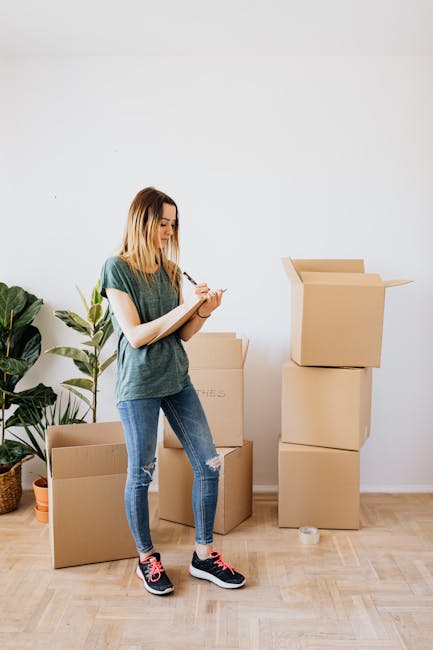 A young woman with long blonde hair, dressed in a casual grey T-shirt and jeans, is standing indoors next to several packed cardboard boxes, some sealed and others open with flaps raised. She is writing on a notepad with a pen, likely making notes related to her home relocation or packing process. Behind her, a large green houseplant adds a natural element to the neutral white-wall setting. In the background, there are additional boxes stacked on the floor, with one partially visible labeled 'CLOTHES.' The scene captures a moment of organisation during packing or a residential move, aligning with the services offered by Man and Van Finsbury for small flat removals and furniture transport as part of comprehensive house removals and relocation logistics.