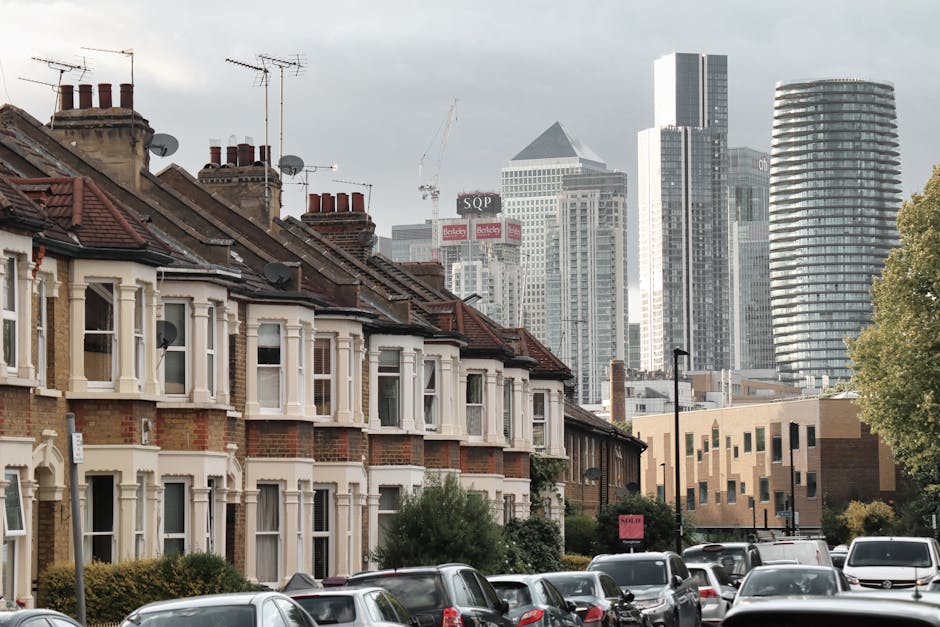 A row of traditional terraced houses with brick facades and white window frames, situated on a residential street with parked cars lining the pavement. The rooftops are equipped with multiple chimneys and satellite dishes, with some rooftops featuring red tiles and others darker materials. Behind the houses, a modern city skyline is visible, including several high-rise office buildings and construction cranes, indicating ongoing development. The scene is captured during daylight with natural lighting and partly cloudy skies. In the foreground, several cars are parked parallel along the street, and a lamppost is visible on the pavement. This image illustrates a typical neighbourhood undergoing an urban transition, relevant to home relocation and furniture transport processes involved in house removals, supported by companies like Man and Van Finsbury, who offer comprehensive packing and moving services near central London.