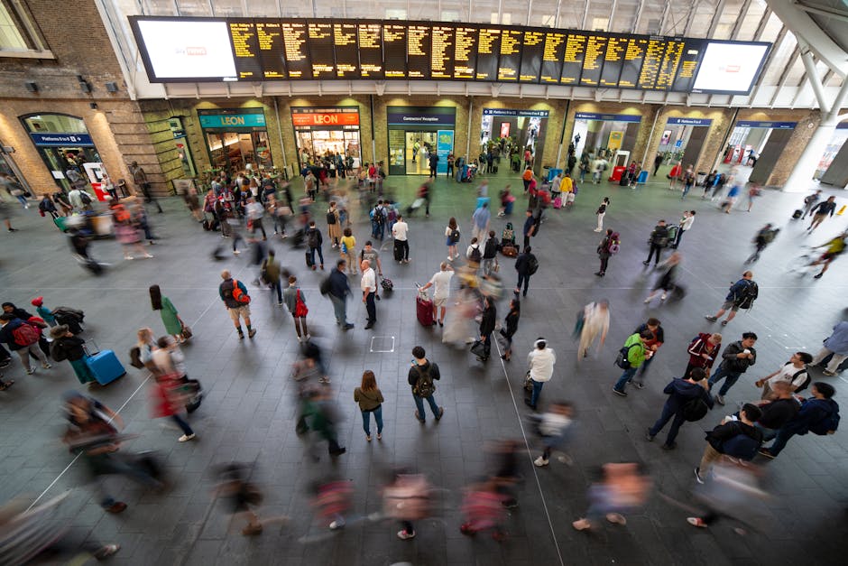 Inside a busy train station with a high, open ceiling and large electronic departure boards displaying train information at the top. The station's interior includes brick walls and glass doors leading to platforms, with crowds of commuters walking, waiting, and carrying luggage across the large, open tiled floor. Some individuals are seen holding or pushing wheeled suitcases, backpacks, or tote bags. Moving figures appear blurred due to motion, while stationary people are more clearly visible. Several passengers are near the station entrances and exits, with some standing in groups or waiting against the walls. The environment is well-lit with natural and artificial lighting. This scene depicts typical busy travel logistics, relevant to house removals or furniture transport planning, as handled by companies like Man and Van Finsbury during home relocations involving strategic packing, loading, and moving process management.