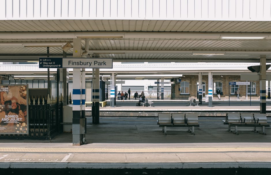 A photograph of Finsbury Park station platform showing several wooden benches placed along the concrete surface and a few passengers sitting and waiting. The station is outdoors with a roof overhead supported by metal columns, and blue and white striped safety features are visible on some infrastructure elements. A sign indicating the station name, 'Finsbury Park,' hangs beneath the roof, and a digital timetable display is mounted on a pillar further down the platform. On the platform edge, a yellow tactile strip runs parallel to the tracks, which are visible in the background along with other passengers standing or walking nearby. To the left side of the image, there is a black fence with a poster advertising 'Romeo and Juliet,' indicating cultural events in the area, and an underground walkway entrance is partially visible. The setting is well-lit with natural daylight, and the scene captures the typical environment where house removals and furniture transport are coordinated during home relocation processes, with a focus on logistical support at busy stations. Man and Van Finsbury occasionally operates within this environment for efficient packing, loading, and transportation of household items.