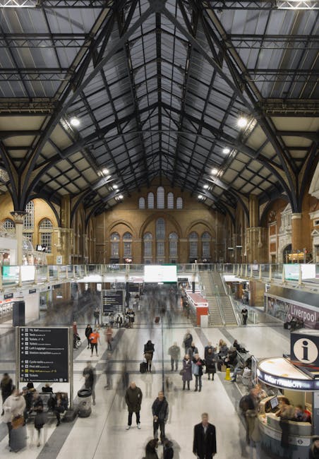 Inside a busy train station with a high, open ceiling and large electronic departure boards displaying train information at the top. The station's interior includes brick walls and glass doors leading to platforms, with crowds of commuters walking, waiting, and carrying luggage across the large, open tiled floor. Some individuals are seen holding or pushing wheeled suitcases, backpacks, or tote bags. Moving figures appear blurred due to motion, while stationary people are more clearly visible. Several passengers are near the station entrances and exits, with some standing in groups or waiting against the walls. The environment is well-lit with natural and artificial lighting. This scene depicts typical busy travel logistics, relevant to house removals or furniture transport planning, as handled by companies like Man and Van Finsbury during home relocations involving strategic packing, loading, and moving process management.