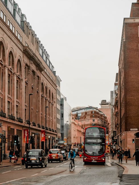 A busy city street scene in Finsbury with multi-storey buildings lining both sides. The buildings feature a mix of brick and modern facades, with retail stores visible at street level and large windows above. Several pedestrians are walking along the sidewalks, some carrying packages or bags. A cyclist is riding in the middle of the street, and multiple cars are parked or moving along the road. A prominent red double-decker bus, displaying route information on the front, is approaching or stopped mid-journey. The street has traffic lights and street signs, with overcast sky overhead. The scene captures an active, urban environment typical for home relocation logistics, where ongoing furniture transport and packing movements could take place. As part of a house removals service, [COMPANY_NAME], such as Man and Van Finsbury, would coordinate loading and unloading of furniture and boxes in this bustling setting, ensuring efficient relocation processes in narrow street conditions.