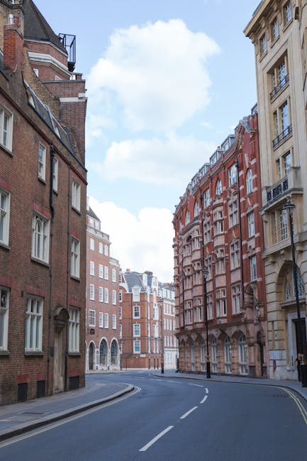 A quiet city street in Finsbury with narrow sidewalks and tall, multi-story residential buildings constructed from brick and stone, featuring large windows and decorative architectural details. The buildings are closely spaced, creating a canyon-like effect, with some featuring small balconies and ornate cornices. The street is paved with asphalt and has white dashed lines indicating traffic lanes. There are black lamp posts along the pavement, and the sky overhead is partly cloudy with patches of blue visible. The scene appears to be captured during daylight hours with natural lighting, reflecting an urban environment suitable for house removals and relocation services in Finsbury. This setting provides context for a home relocation or furniture transport process happening nearby, possibly involving a moving van or crew working in the vicinity, although no vehicles or people are visible in this particular image.