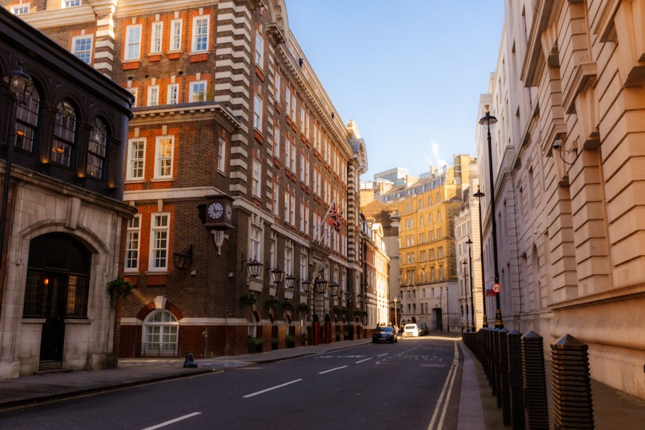 A busy city street scene in Finsbury with multi-storey buildings lining both sides. The buildings feature a mix of brick and modern facades, with retail stores visible at street level and large windows above. Several pedestrians are walking along the sidewalks, some carrying packages or bags. A cyclist is riding in the middle of the street, and multiple cars are parked or moving along the road. A prominent red double-decker bus, displaying route information on the front, is approaching or stopped mid-journey. The street has traffic lights and street signs, with overcast sky overhead. The scene captures an active, urban environment typical for home relocation logistics, where ongoing furniture transport and packing movements could take place. As part of a house removals service, [COMPANY_NAME], such as Man and Van Finsbury, would coordinate loading and unloading of furniture and boxes in this bustling setting, ensuring efficient relocation processes in narrow street conditions.