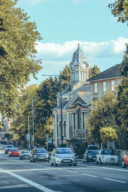 A street scene in Finsbury showing a busy road with multiple cars moving in both directions, parked along the curb, and a sidewalk lined with mature trees. In the background, there is a historic building with a clock tower featuring a conical roof, decorative mouldings, and a clock face visible on both sides. The building appears to be part of a church or civic structure, surrounded by residential and commercial buildings with various roof types. The area is well-lit with natural daylight, and the scene captures the urban environment typical of a house removal or furniture transport process, with parking spaces and street lighting indicating a central area suitable for local removals by [COMPANY_NAME] as referenced on the Finsbury page about same-day moves.