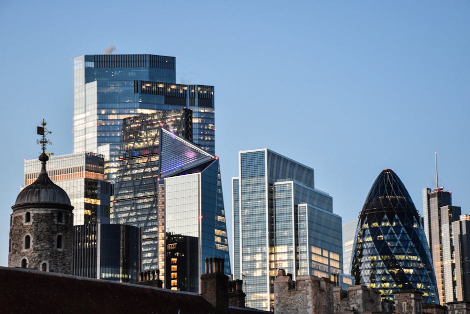 A view of a modern city skyline featuring several tall, reflective glass skyscrapers with illuminated windows, including the distinctive Gherkin building with its rounded, diamond-patterned exterior, and the topped-off tower with a large communications antenna on the right. In the foreground, there is a historic stone building with a rounded turret and a weather vane on top, contrasting with the contemporary architecture behind it. The scene is set during early evening or dusk, with the sky clear and a hint of remaining daylight. This urban landscape showcases the blending of historic and modern structures typical in areas like Finsbury. For house removals or furniture transport, loading and unloading activities would take place near such buildings, often involving the careful handling of furniture and boxes, possibly wrapped in blankets or plastic for protection during a home relocation or moving service, as provided by companies like Man and Van Finsbury.