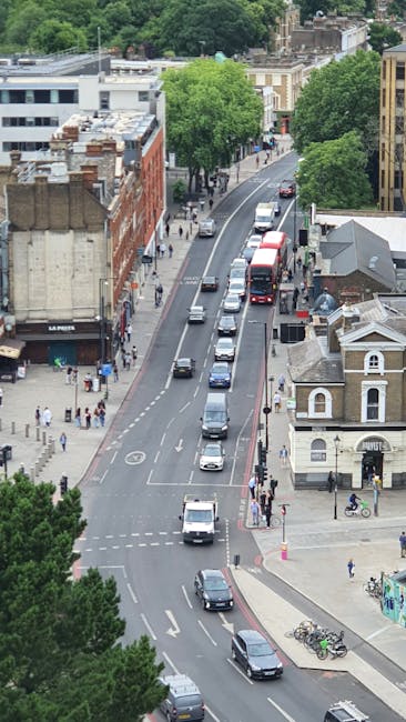 An aerial view of Finsbury town centre showing a narrow, one-way street with several parked and moving vehicles, including cars, a red double-decker bus, and delivery vans. The street is lined with a mix of residential and commercial buildings, including a white house with a small garden and a brick building with a sign for a retail store. Pedestrians are visible walking along the sidewalks, some waiting at crosswalks, while others are engaged in outdoor activities. Large, leafy green trees provide partial shade along the street, and visible parking bays contain bicycles secured to bike racks. The environment appears urban with a combination of older and modern architecture, and the scene is well-lit with natural daylight. This setting depicts typical city traffic and pedestrian movement, relevant to the context of home relocation and logistical planning for removals conducted by Man and Van Finsbury, especially in a busy area like Finsbury Common during a weekend. The image captures both the street traffic flow and surrounding urban infrastructure, pertinent to furniture transport and moving logistics in a residential and commercial neighbourhood.