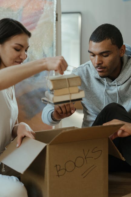 A woman and a man are inside a home, engaging in a packing or moving process. The woman is holding a cardboard box labeled 'BOOKS' on its side, while the man is lifting a small stack of books wrapped with a rubber band, preparing to place them into the box. The background shows a blurred view of a doorway and a painted wall, indicating an interior space. The scene involves furniture and packing materials such as cardboard boxes and books, illustrating home relocation preparations. This image reflects the packing stages of a house move, showcasing the careful handling and organization of items typical in professional removals services like those offered by Man and Van Finsbury, as featured on their website's removals service page.